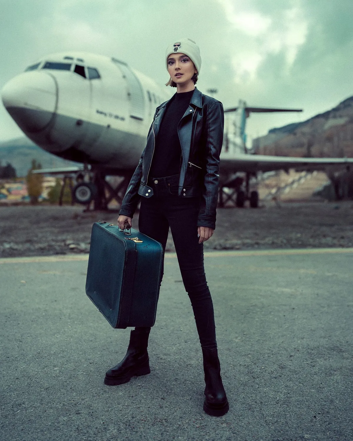 A woman in a leather jacket holding a suitcase near an airplane.