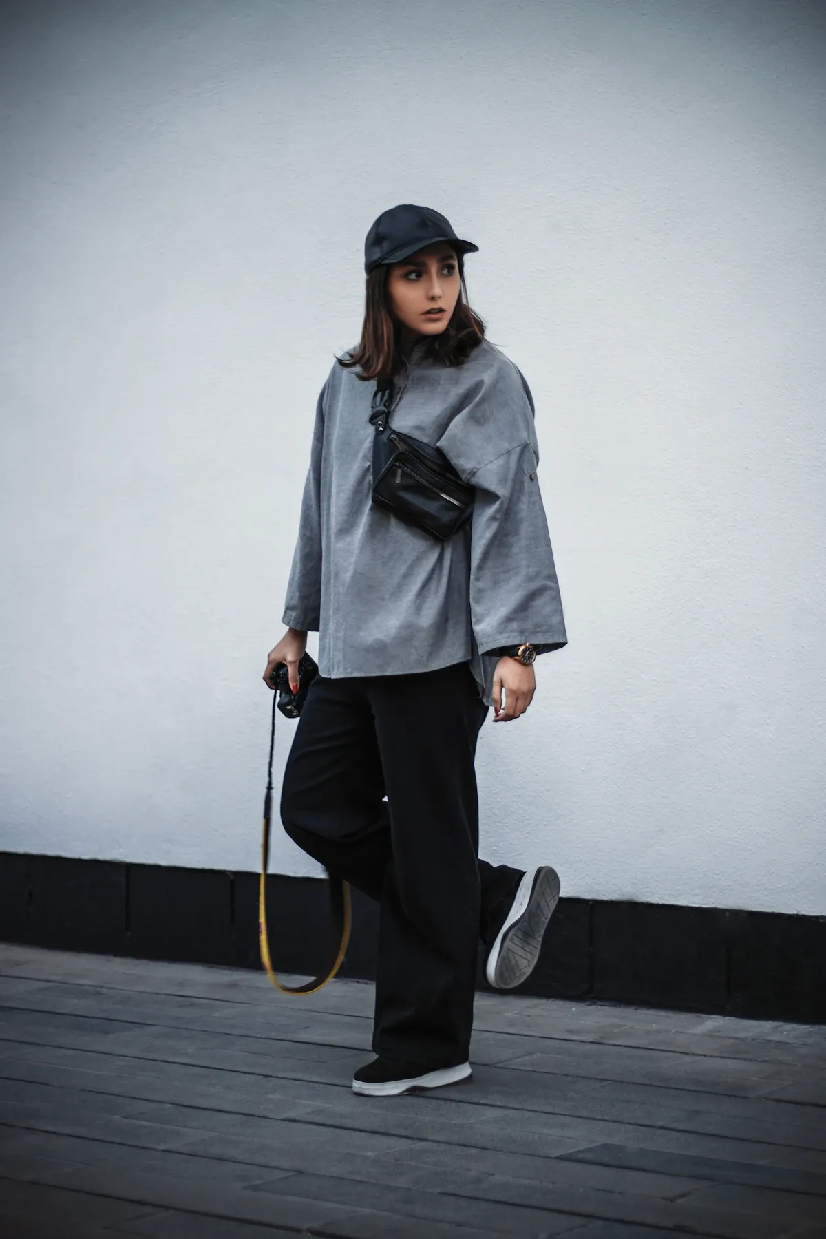 A woman in a grey top, dark trousers and a dark cap beside a pale wall.