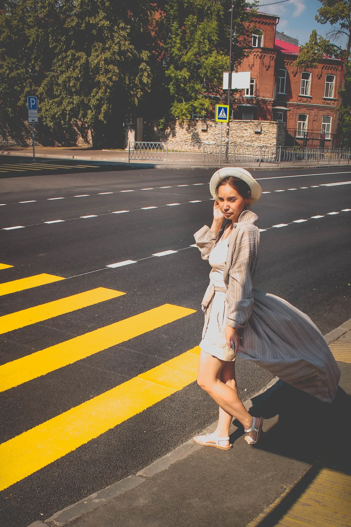 A woman in a pale dress, striped outer layer and hat beside a marked city crossing.