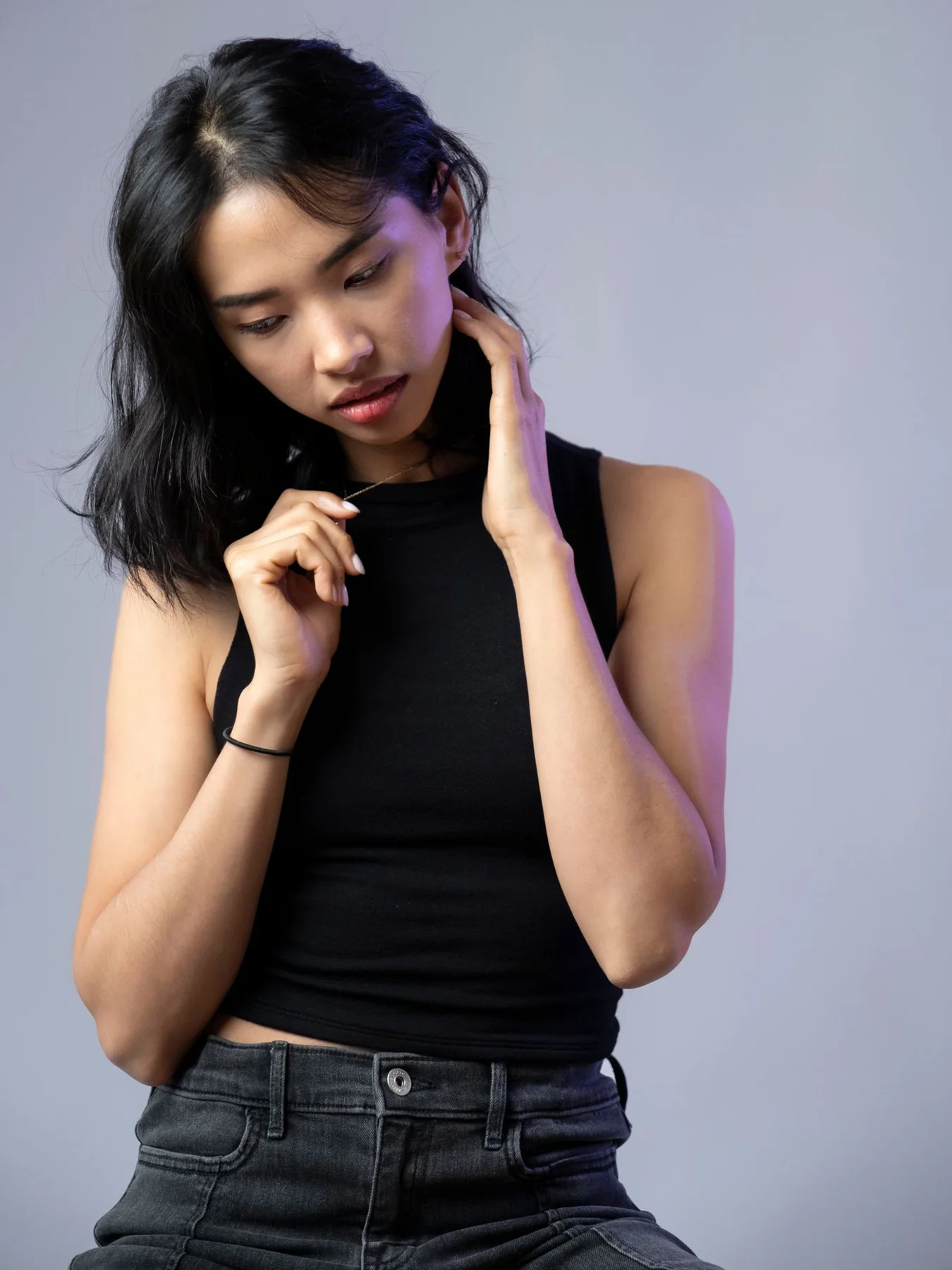 A woman in a dark sleeveless top and jeans against a grey background.