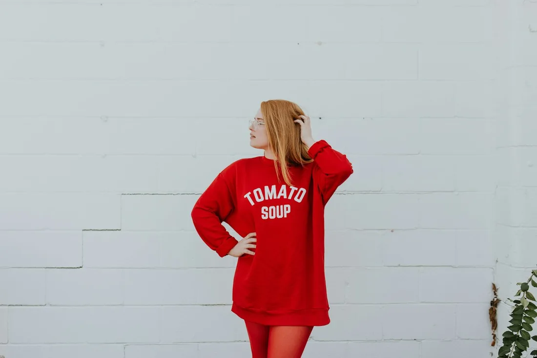 A woman in an oversized red graphic sweatshirt beside a pale wall.