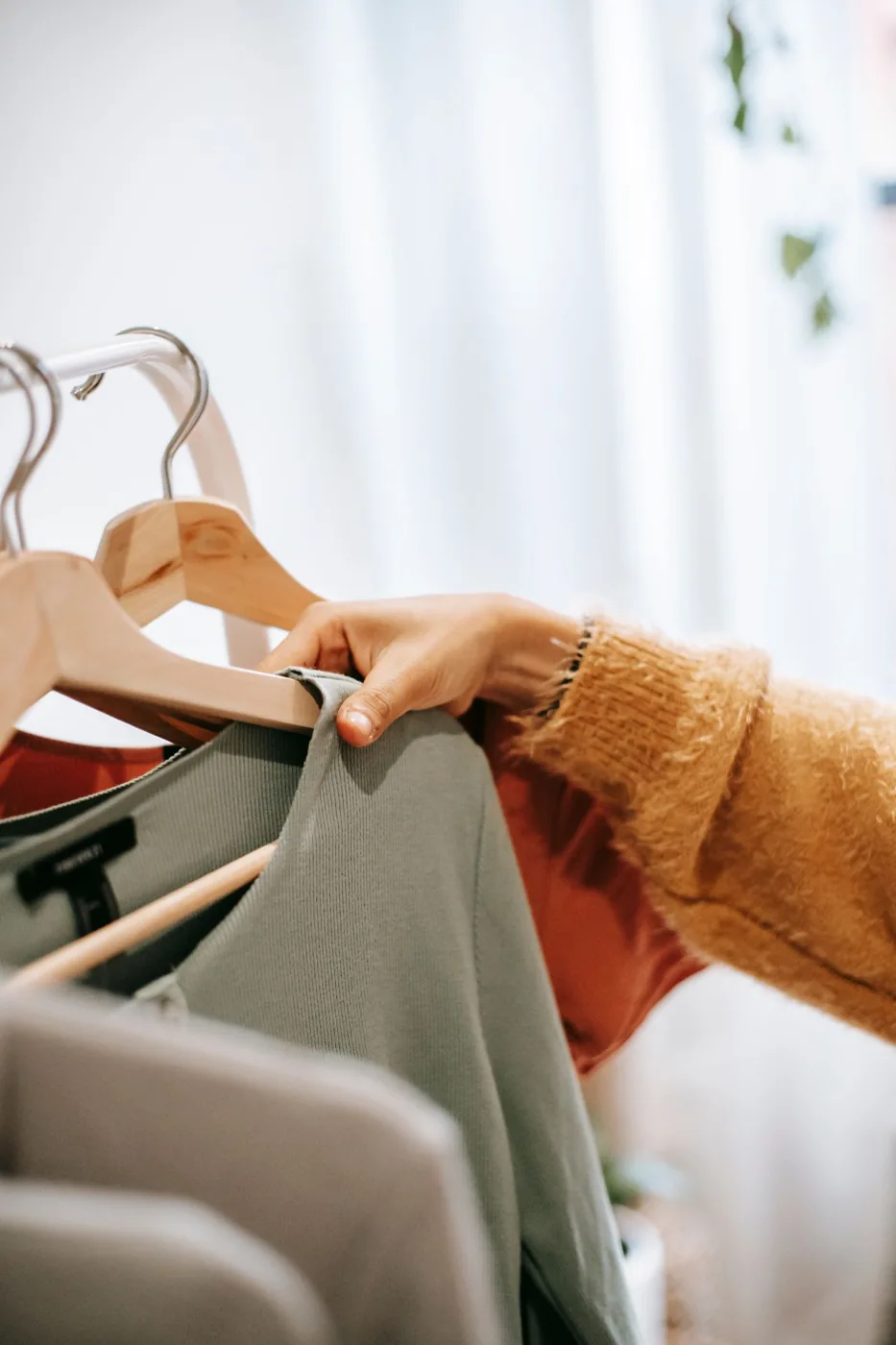 A hand among garments on wooden hangers beside a bright window.