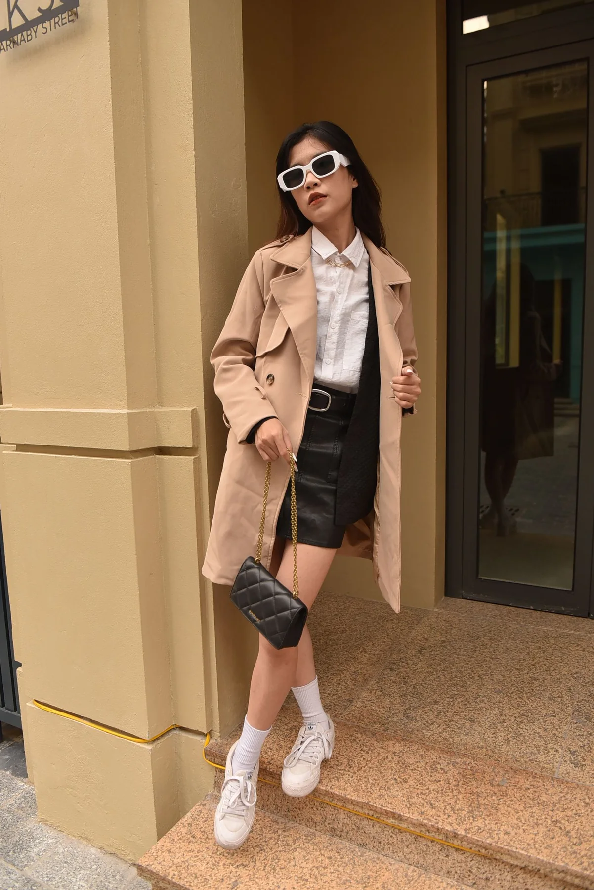 A woman in sunglasses and a pale coat beside a doorway.