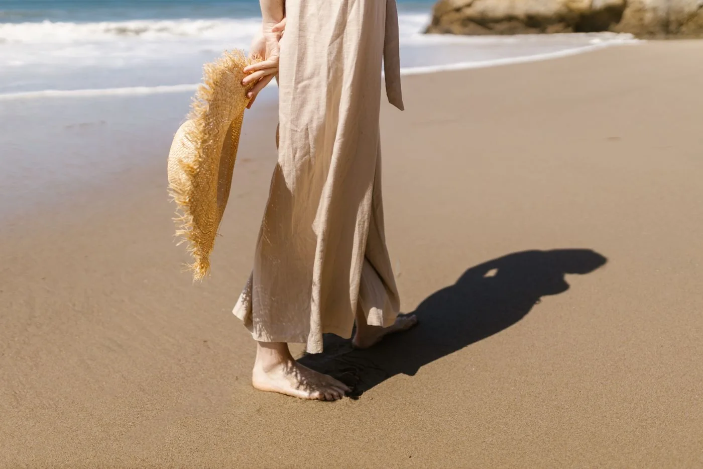 A person in pale trousers holding a straw hat beside the sea.