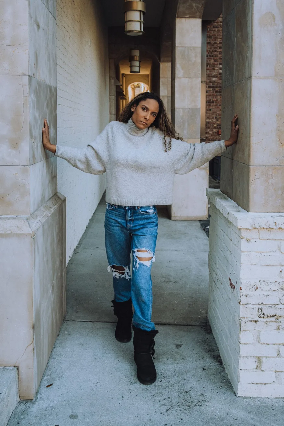 A woman in a pale sweater and jeans between stone walls.