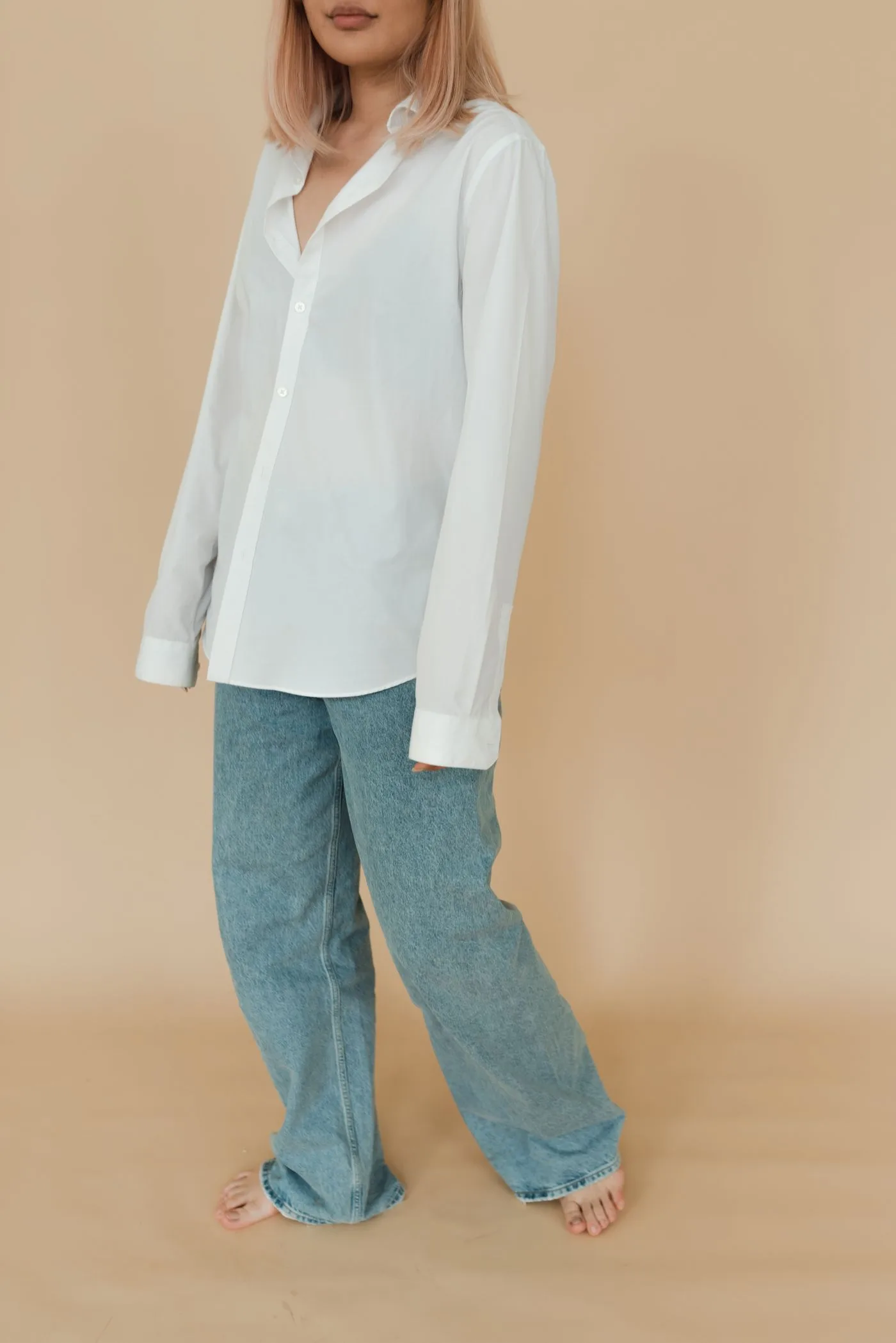 A person in a loose pale shirt and jeans against a pale wall.
