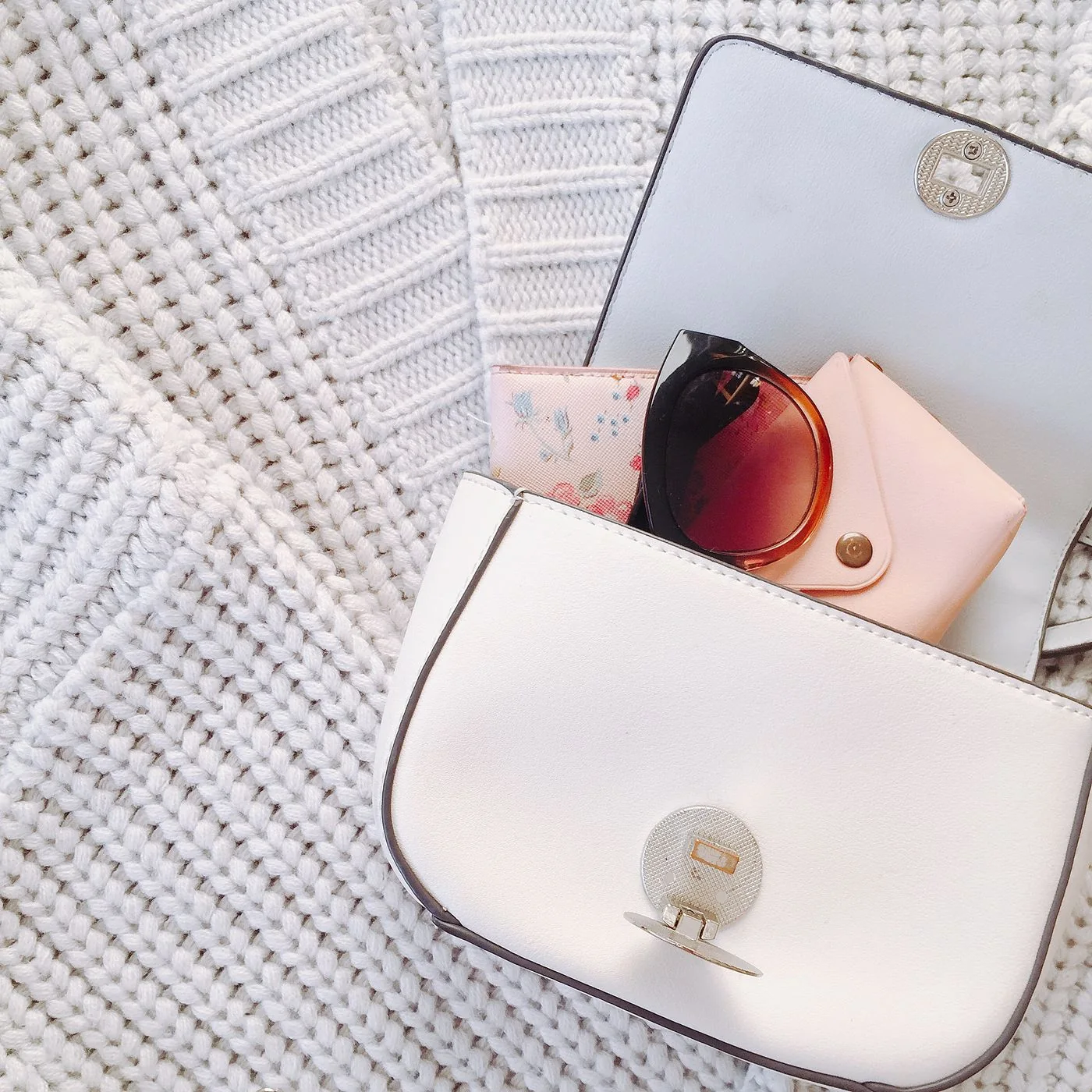 A pale handbag beside sunglasses and small accessories on a pale knit surface.