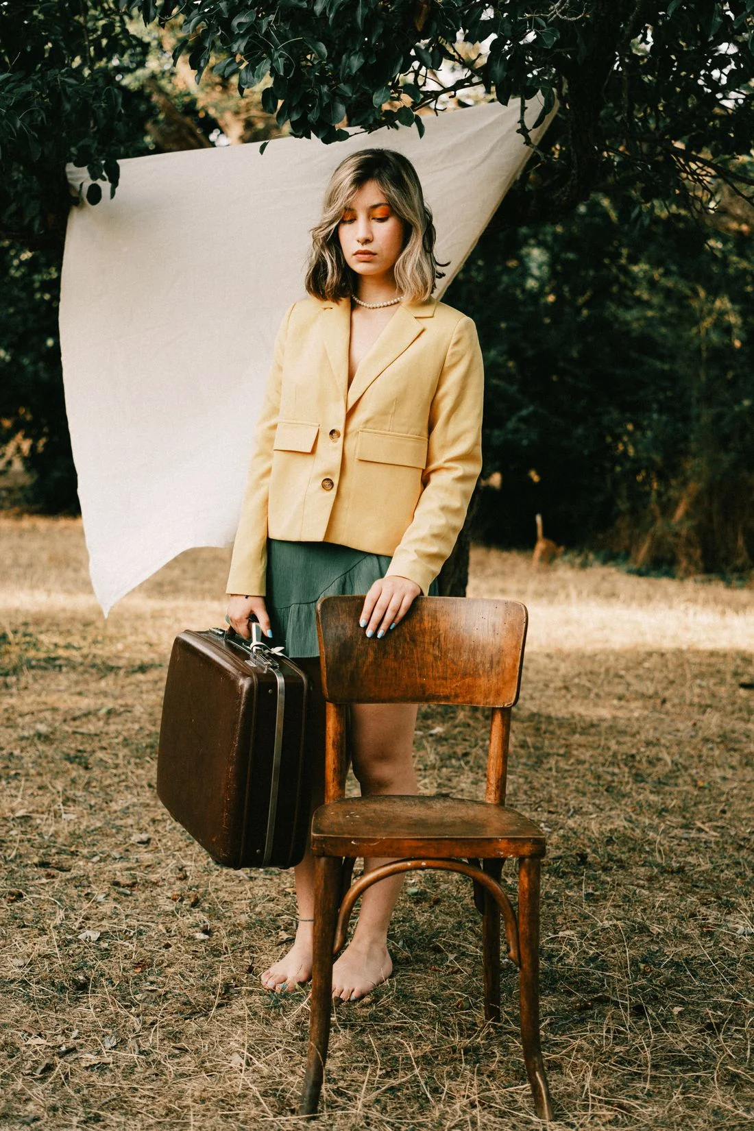 A woman in a pale yellow jacket carrying a dark structured case beside a wooden chair.