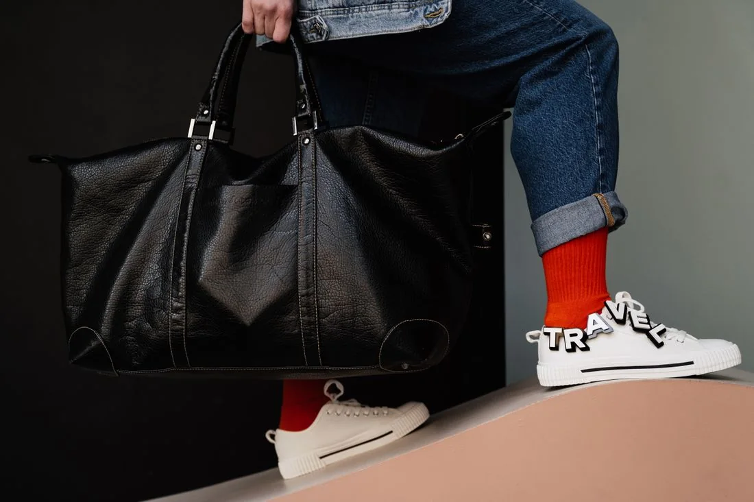 A person holding a dark travel bag beside pale sneakers and rolled trouser hems.