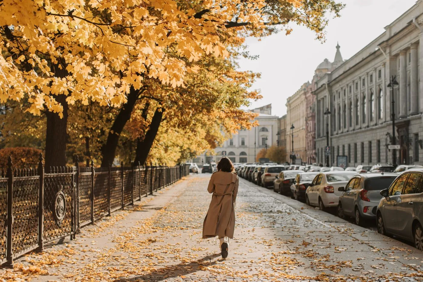 A woman in a long pale outer layer on a leaf-covered city pavement.
