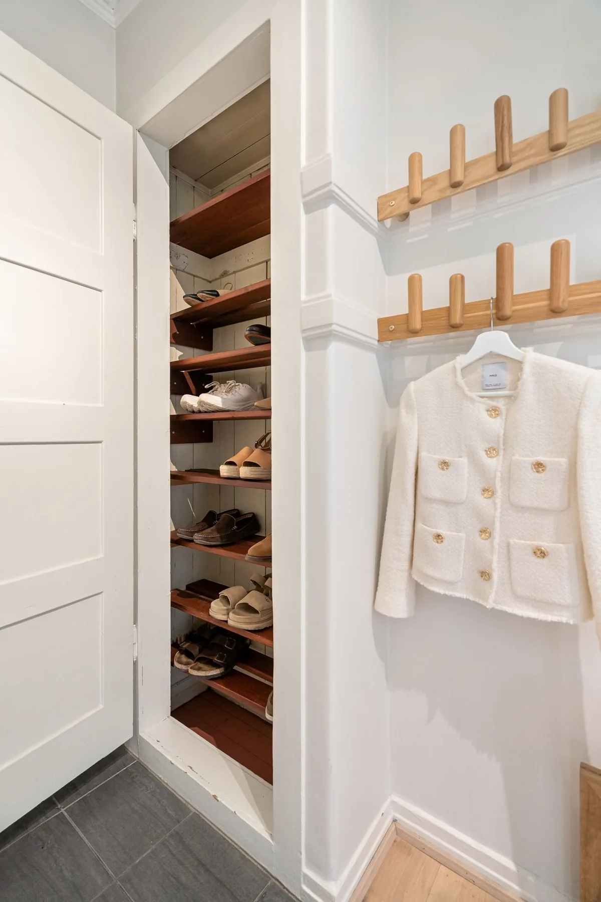 A white jacket on a hanger suspended from wooden wall hooks beside an open shoe cupboard.
