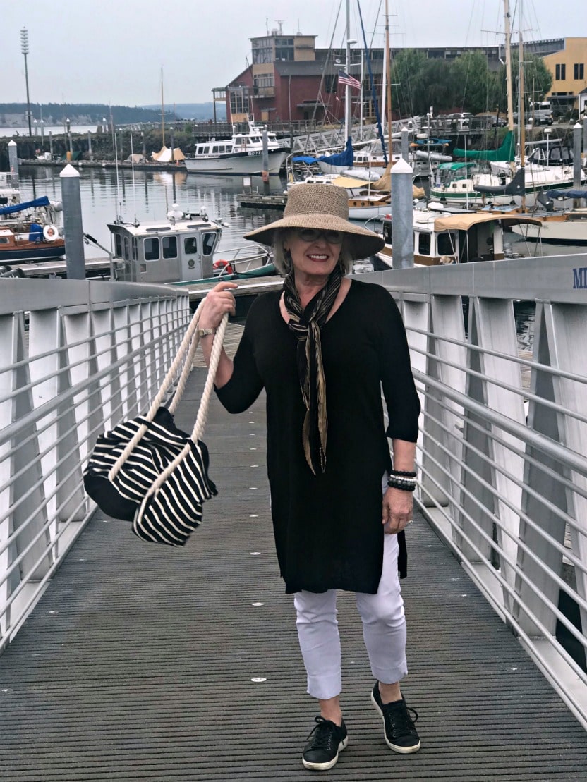 Jennifer Connolly of A Well Styled Life wearing long black tunic over white stretch pants with beige hat and black sneakers on the dock at Port Townsend, WA