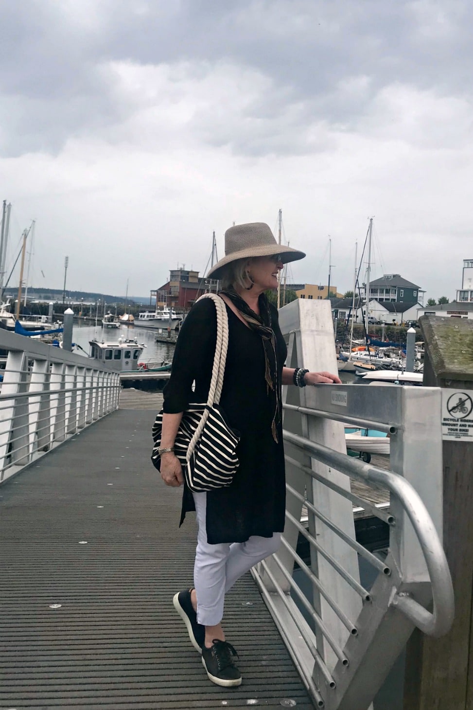 Jennifer Connolly of A Well Styled Life wearing black tunic and white pants with black sneakers and beige hat on the dock at Port Townsend