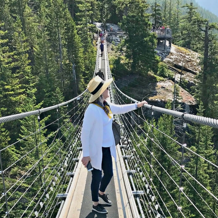 Jennifer Connolly of A Well Styled Life wearing straw hat, white sweater set and deep blue jeans on suspension bridge