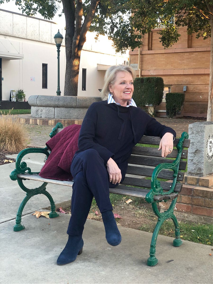 over 50 fashion blogger Jennifer of a well styled life sitting on park bench weaing blue sweater, blue pants and blue boots by Talbots