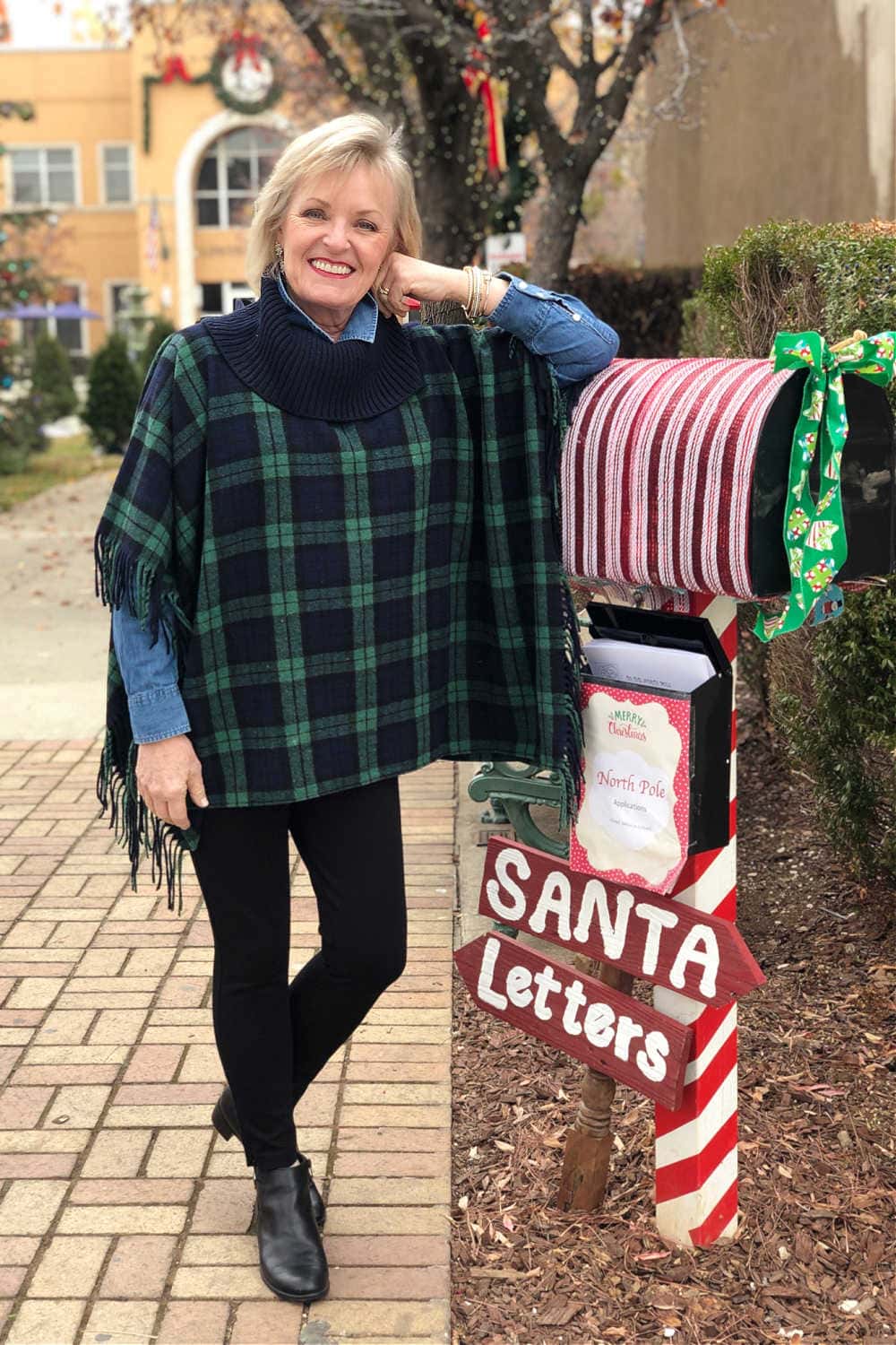 over 50 fashion blogger Jennifer from a well styled life wearing talbots soho leggings and blackwatch plaid poncho leaning on santa's mailbox