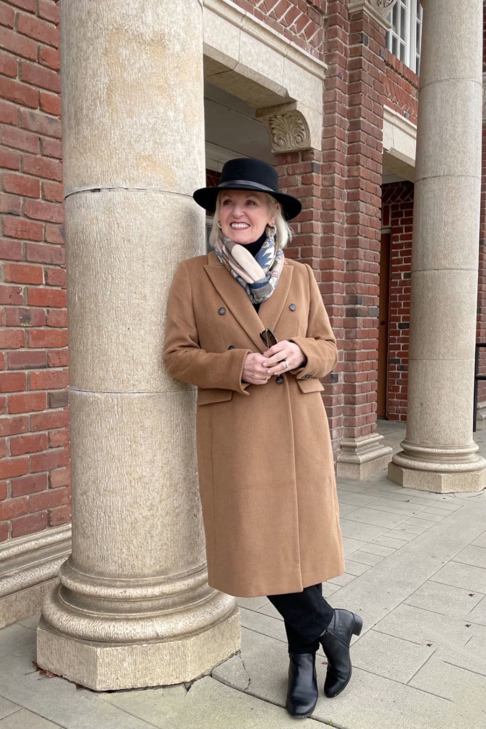 woman leaning against column wearing camel coat and black hat
