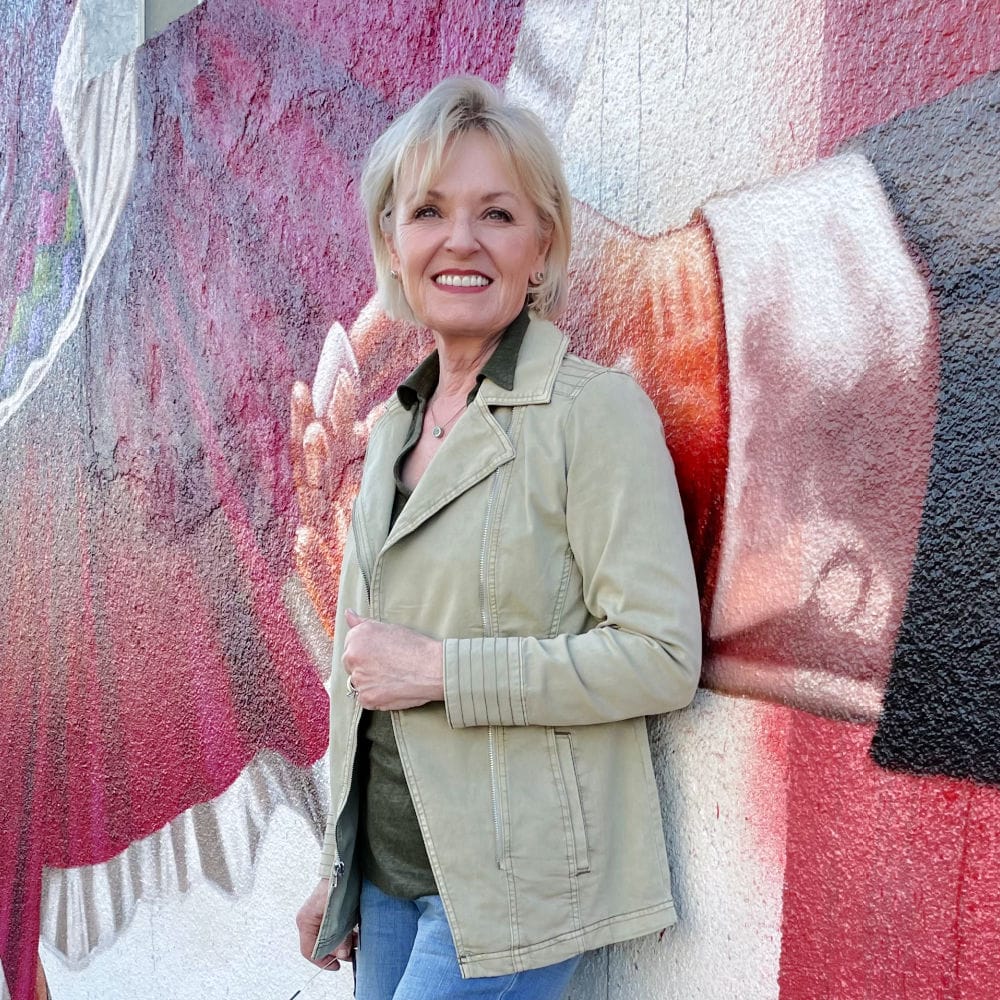 over 50 blogger wearing green jacket and blue jeans leaning against a wall mural