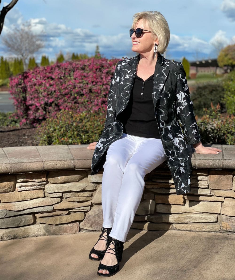 woman sitting on rock wall wearing white jeans, black top and white and black jacket