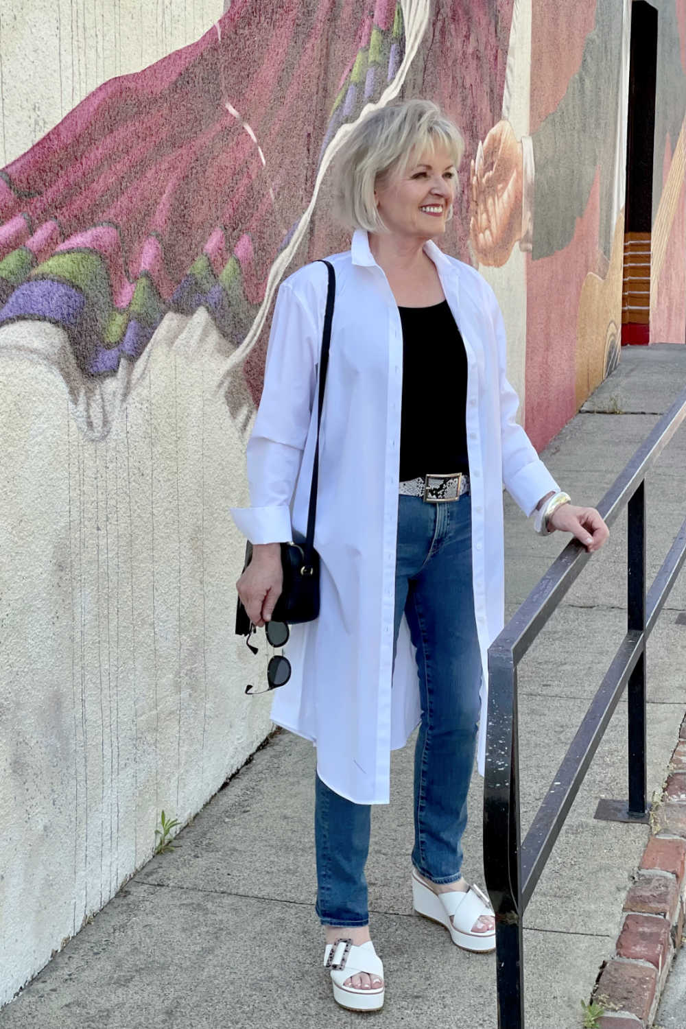 WOMAN IN FRONT OF MURAL WEARING WHITE DUSTER AND BLUE JEANS WITH SNAKEPRINT BELT