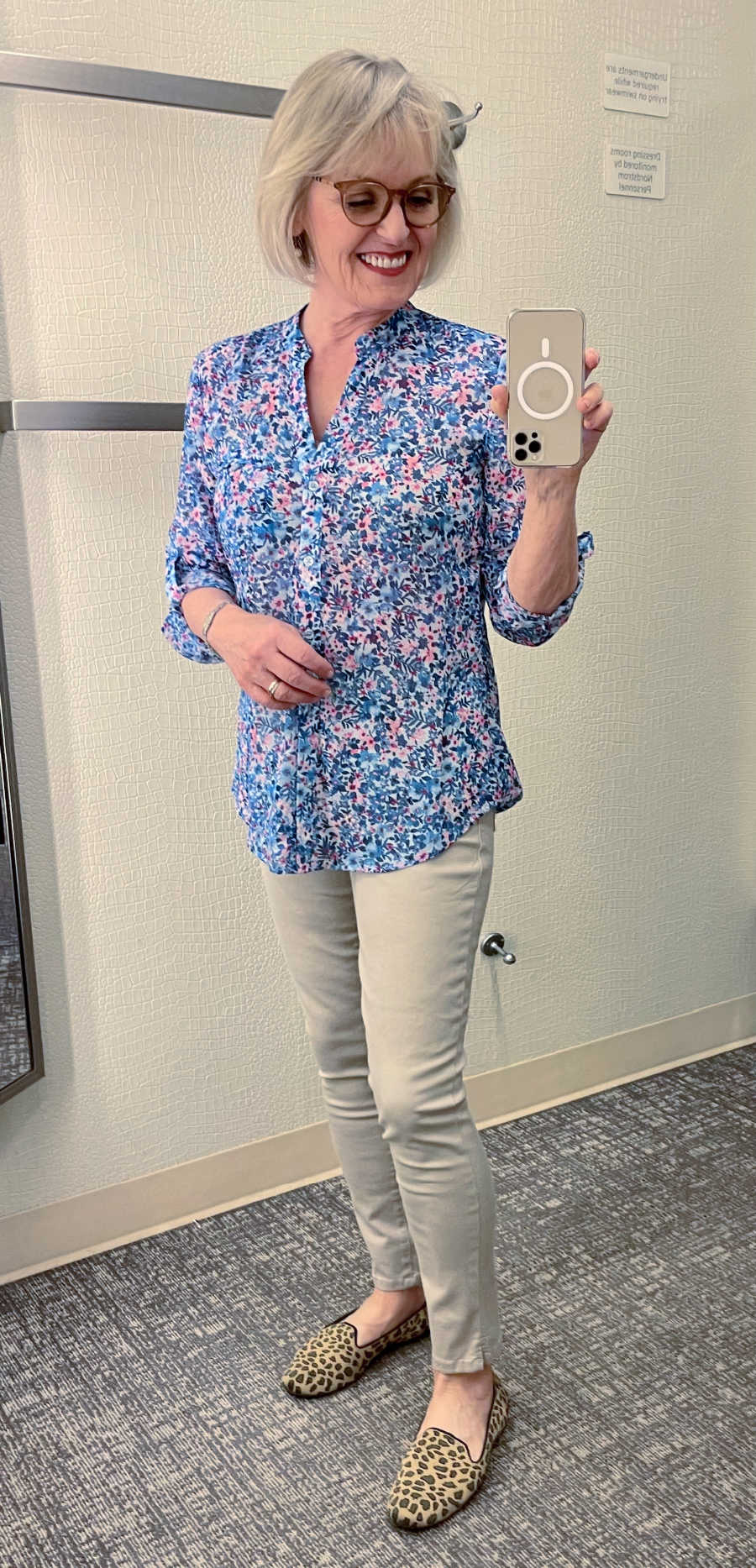 blonde woman taking picture in mirror of her wearing floral blouse and tan jeans