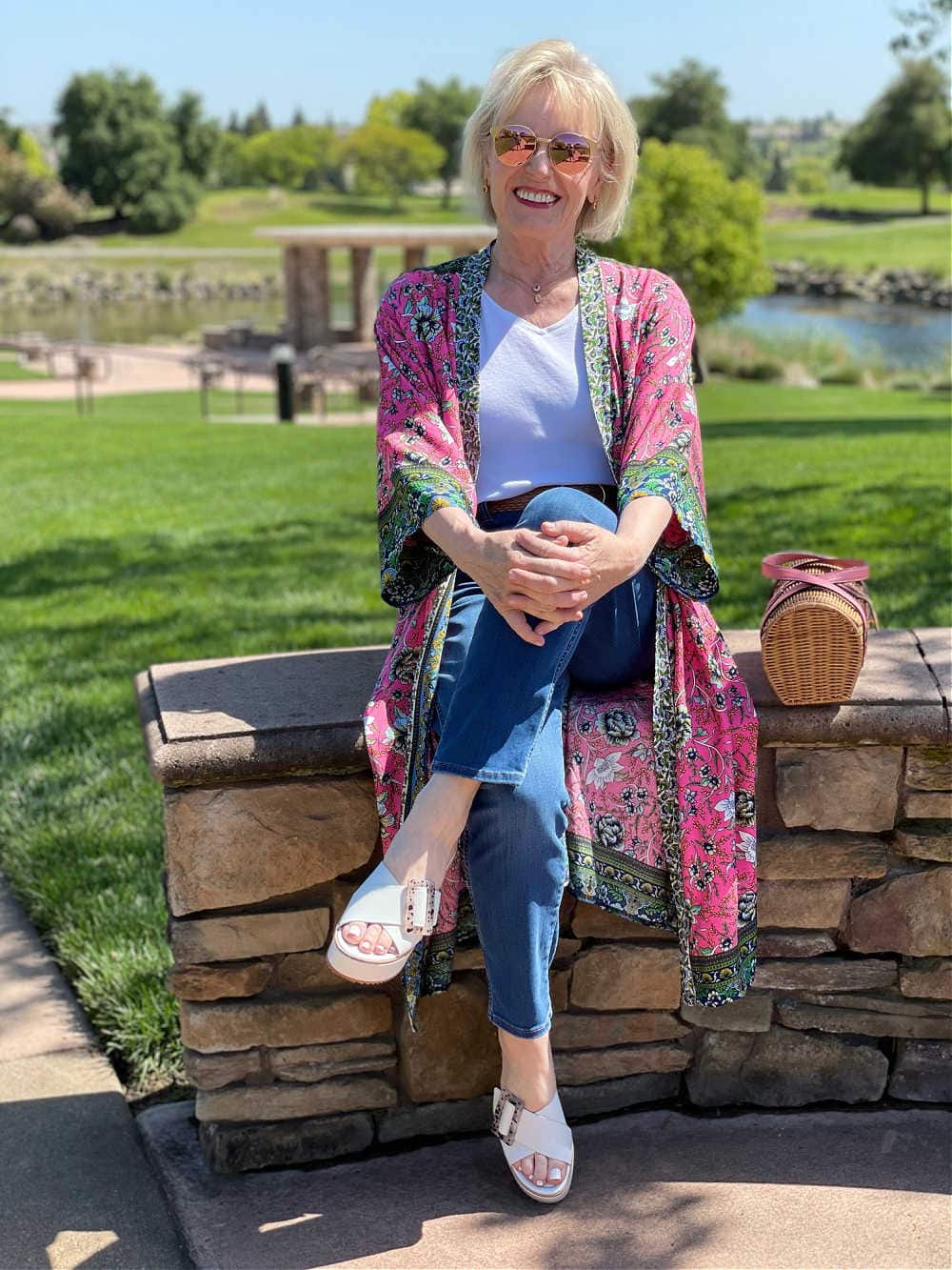 woman sitting on low wall wearing blue jeans and pink kimono
