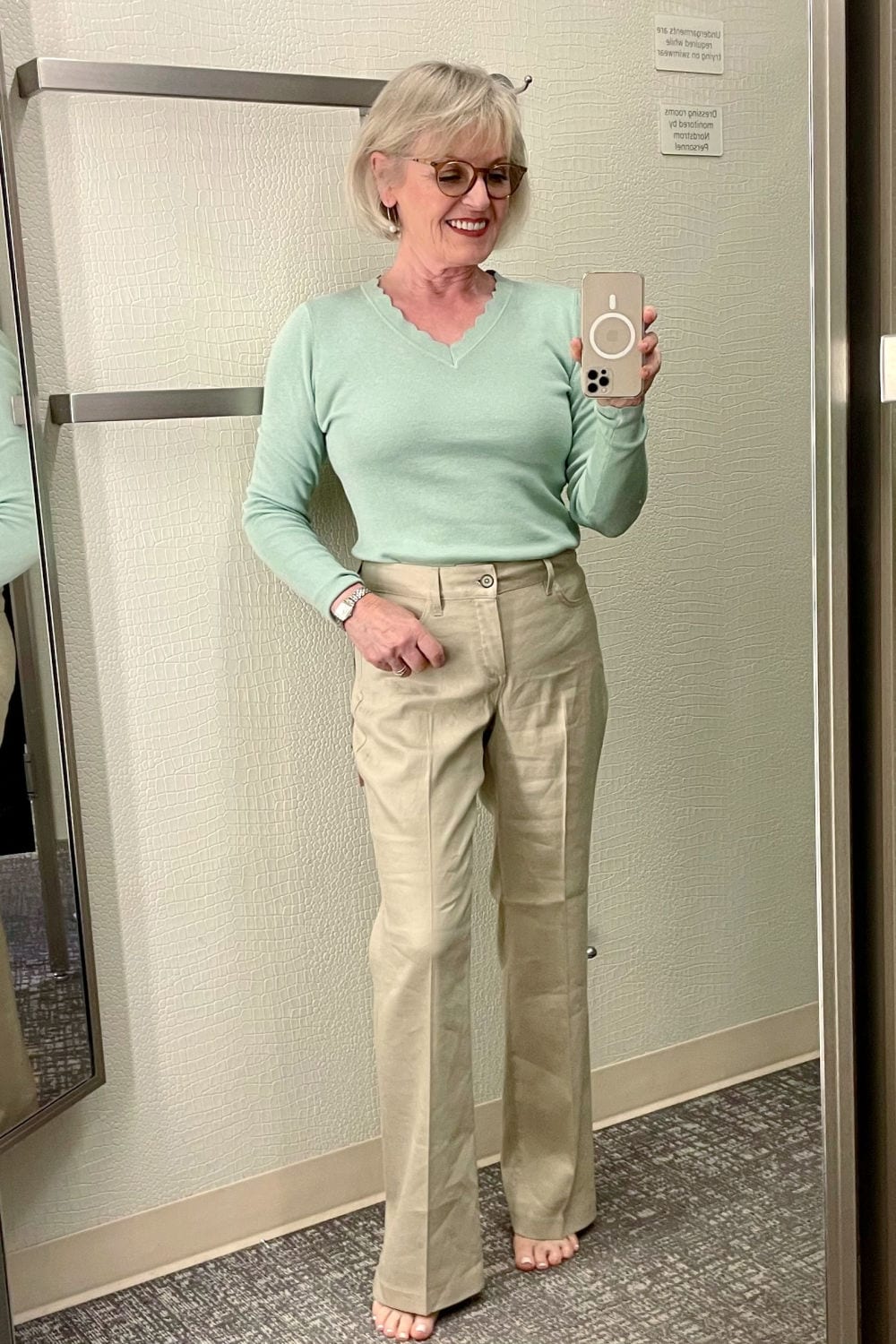 blonde woman in dressing room trying on green sweater and beige linen pants