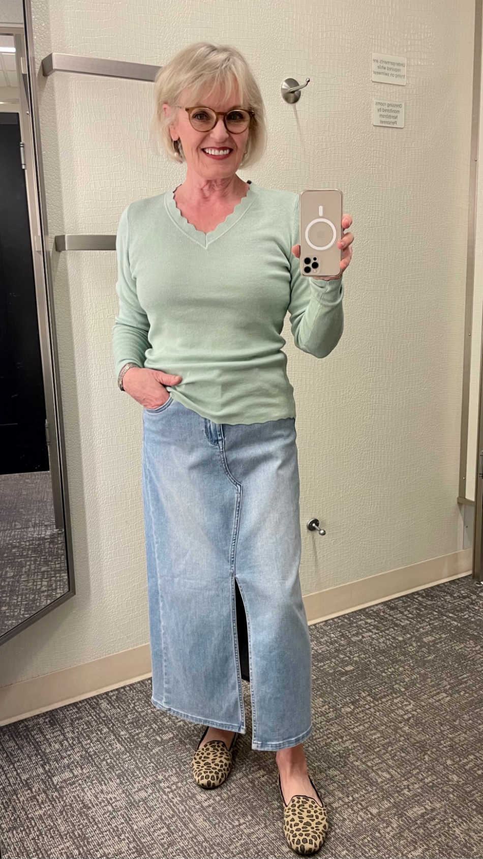 women trying on denim skirt and green sweater in dressing room mirror