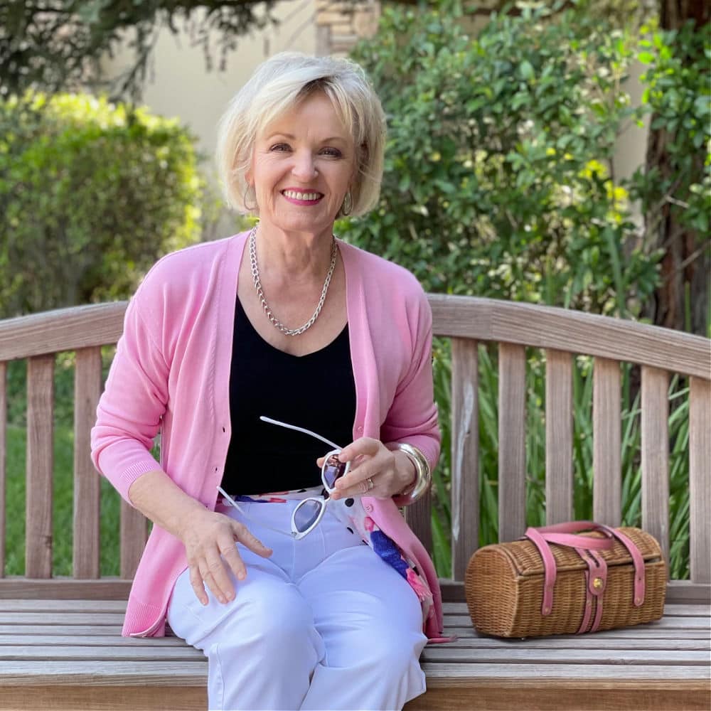 blond woman sitting on bench wearing pin sweater, black tank and white pants