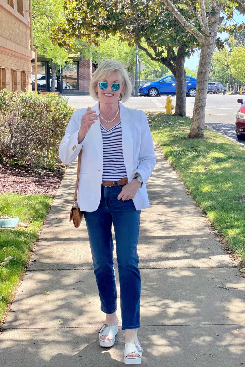 woman walking towards you on sidewalk with mirror sunglasses, white blazer and blue jeans