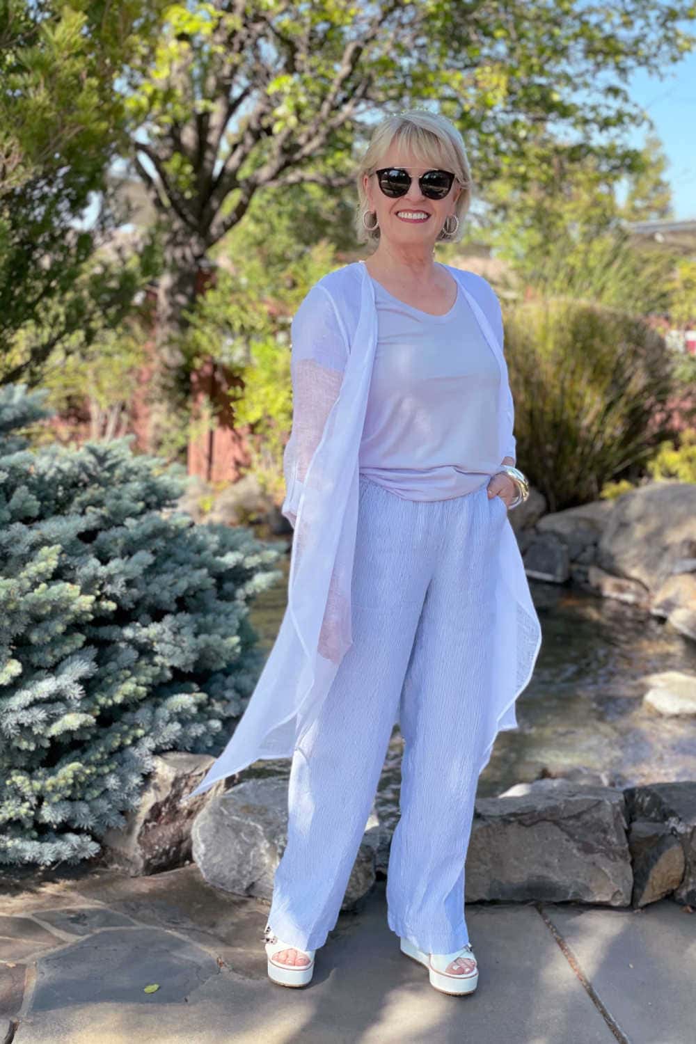 woman standing in front of pond wearing white gauze duster with striped linen trousers