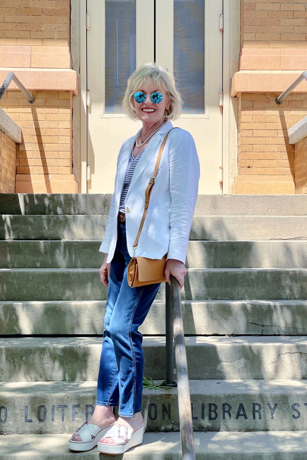 blonde woman leaning on railing os strair wearing white blazer and blue jeans