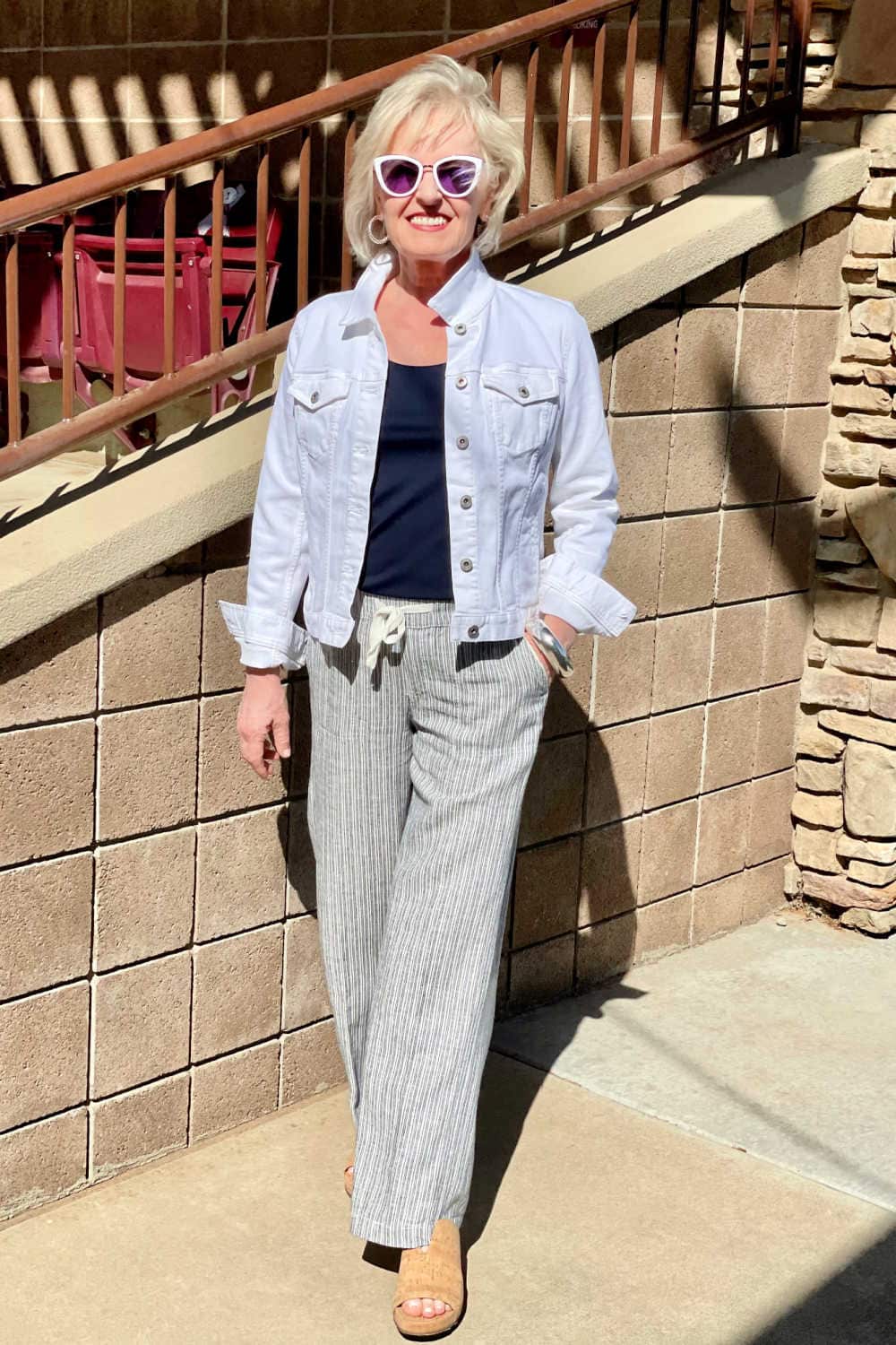 woman walking toward the camera wearing mirrored sunglasses, white jacket, striped pants and cork shoes