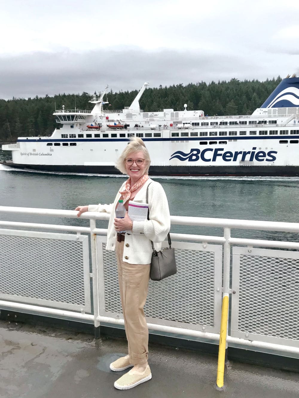 blonde woman standing on dock of ferry wearing cream cardigan, beige cargo pants and cinnamon colored tee shirt
