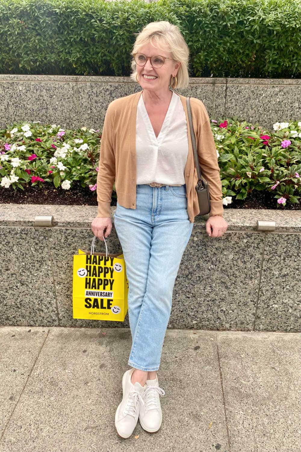 blonde woman wearing camel colored cardigan, blue jeans, metallic sneakers leaning against planter