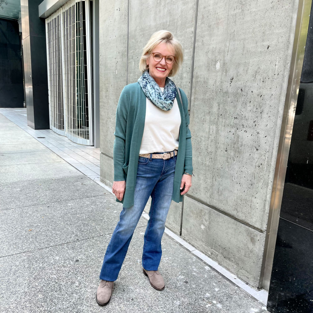 blonde woman standing on sidewalk wearing teal sweater, blue denim and taupe boots