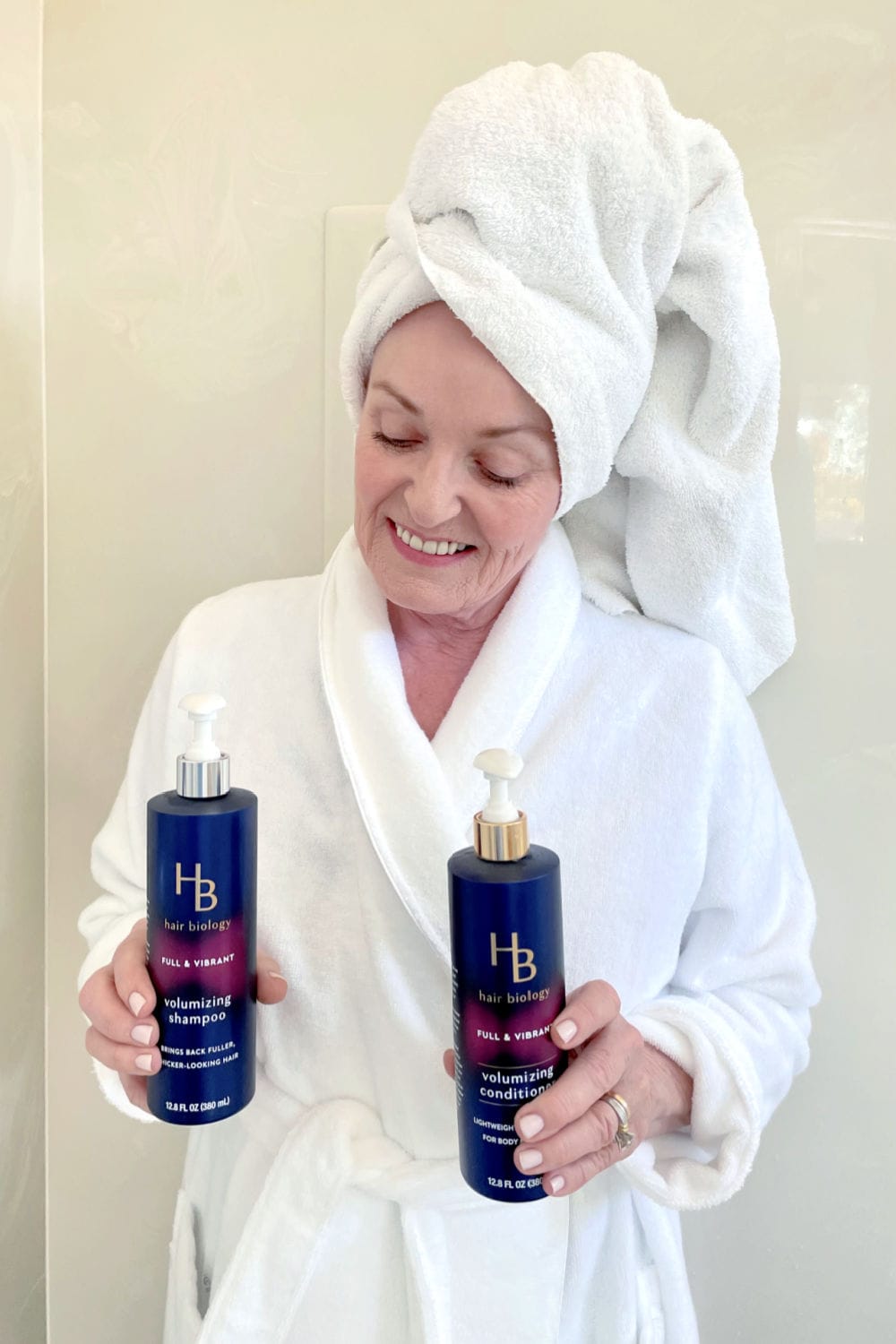 WOMAN IN WHITE ROBE STANDING IN SHOWER HOLDING HAIR BILOGY SHAMPOO AND CONDITIONER