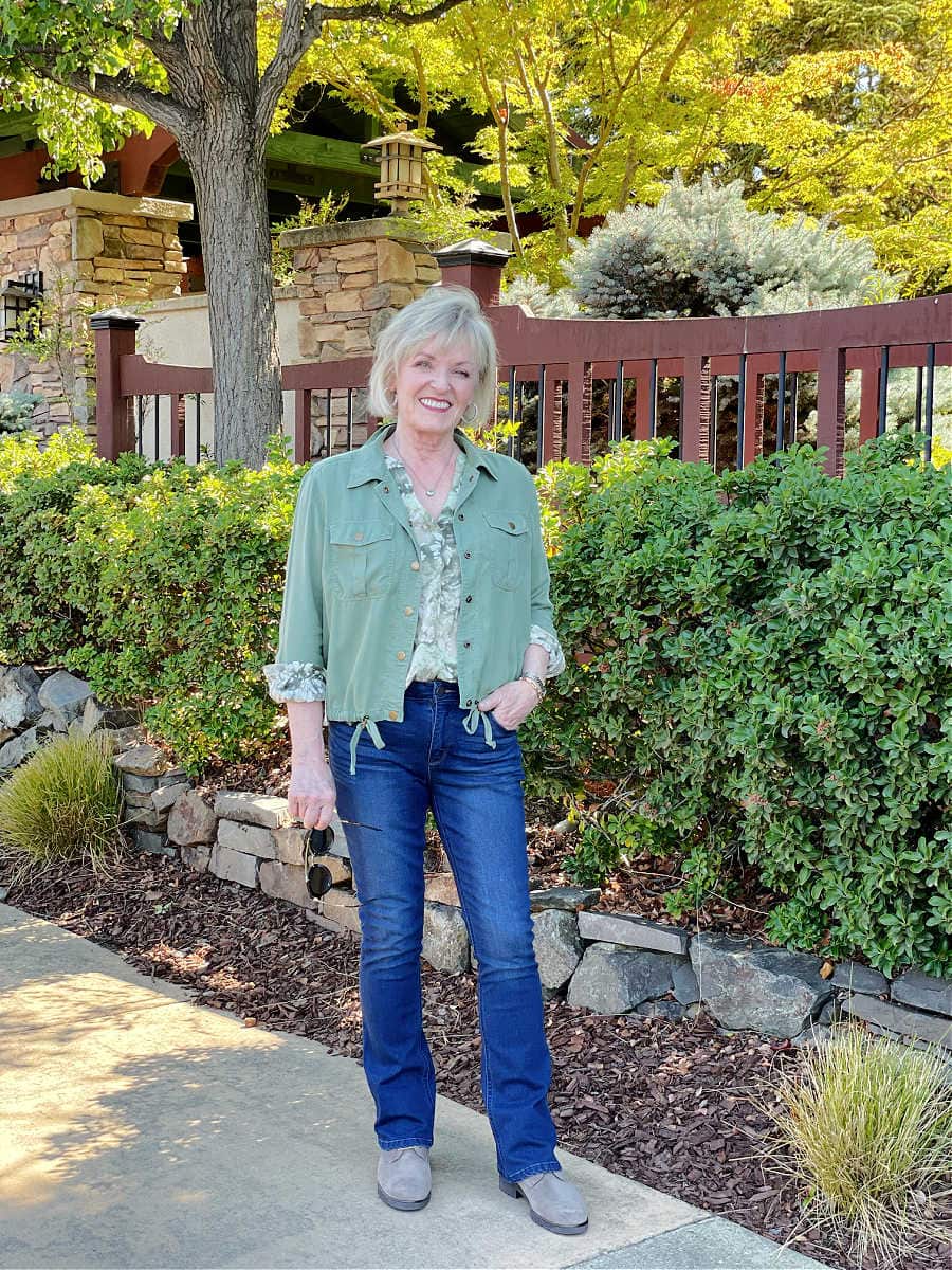 woman wearing floral blouse and lightweight fall utility jacket with jeans and booties standing in front of fence