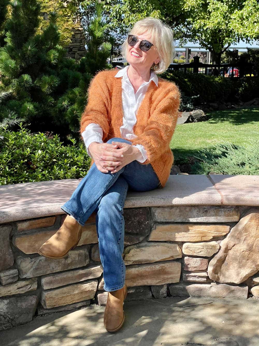 bolnd woman sitting on wall wearing rust colored cardigan and blue jeans