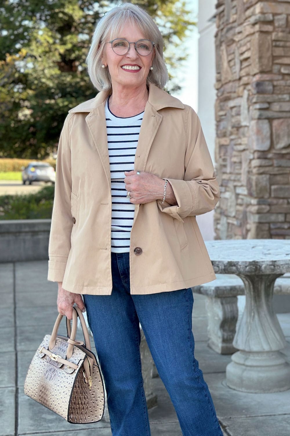 Short trench coat in a soft neutral styled with striped top and satchel bag on woman over 50.