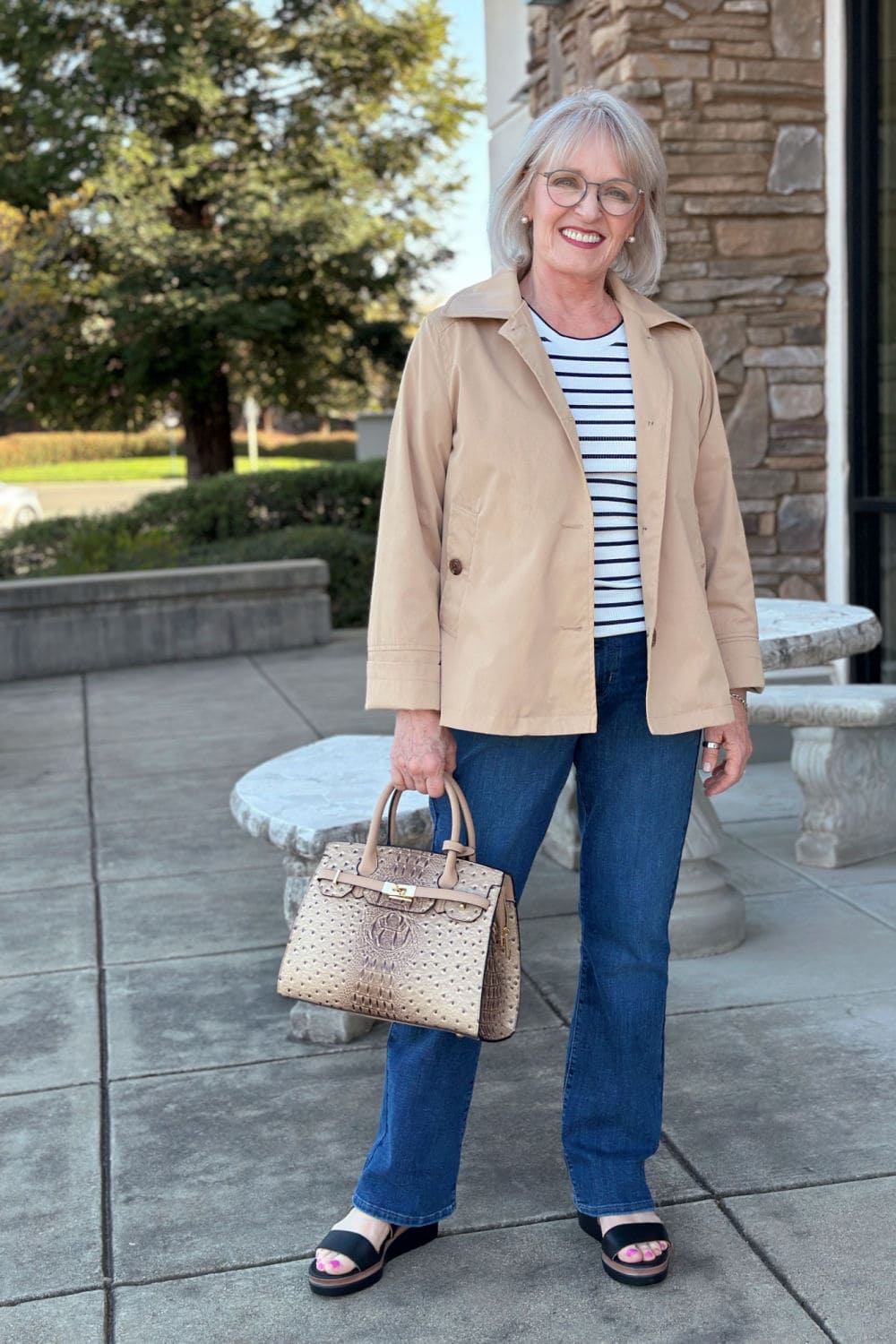 Woman over 50 in a tan short A-line raincoat with dark bootcut jeans and striped top.