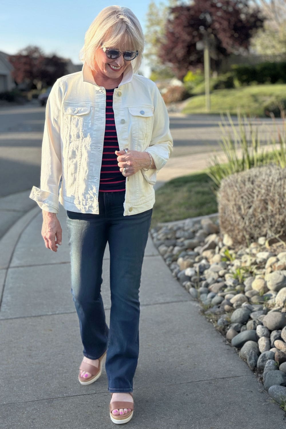 Woman over 50 wearing an ivory denim jacket with bootcut jeans and striped top, styled with wedge sandals.