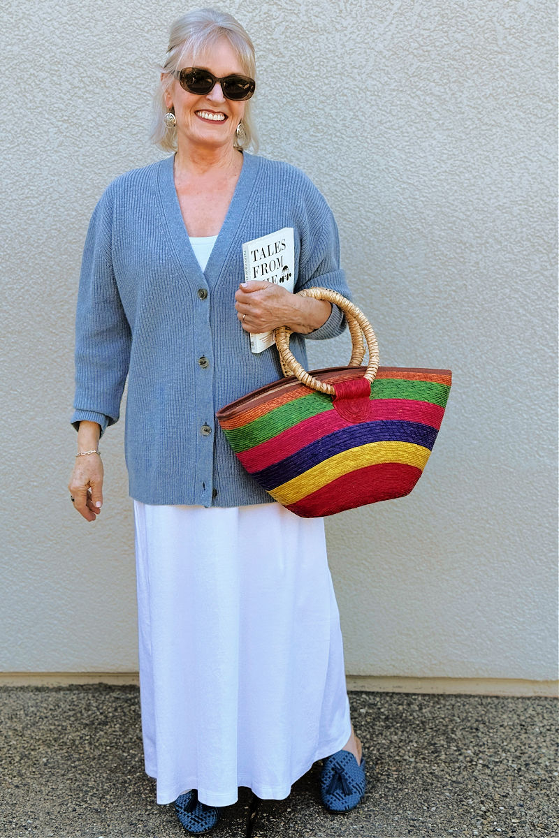 Jennifer of A Well Styled Life wearing a blue ribbed cardigan, white maxi skirt, and a colorful straw tote bag for a spring neutral look.