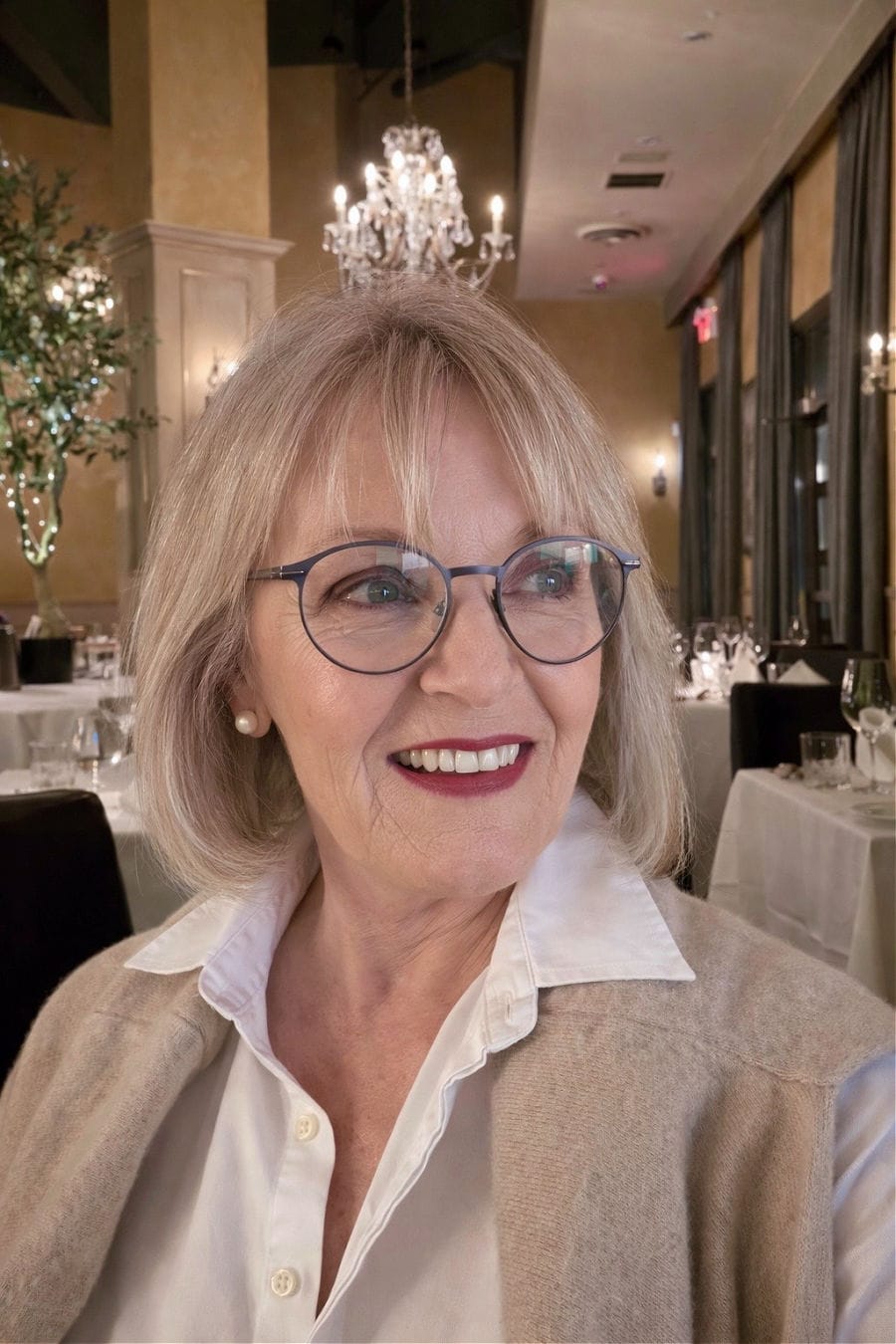 A polished look featuring Jennifer in a white button-down shirt and a tan cashmere cardigan, styled with classic white pearl stud earrings in a restaurant setting.