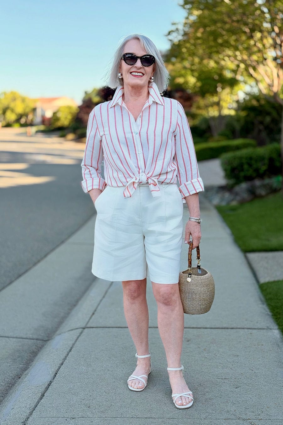 Jennifer Connolly wearing white linen Bermuda shorts with a red and white striped tie-front shirt, white sandals, and a woven bag
